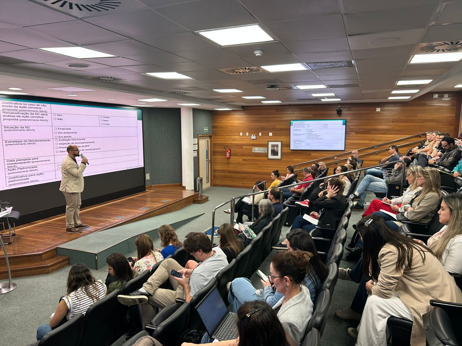 Foto de uma apresentação técnica em auditório. Um palestrante em pé, de blazer bege, segura um microfone e fala diante de uma grande tela que exibe um formulário projetado. O público, composto por profissionais atentos e com anotações ou notebooks, ocupa as poltronas em fileiras. O ambiente é moderno, com paredes de madeira e iluminação clara.