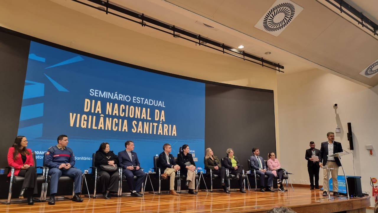 A imagem mostra um auditório durante o Seminário Estadual – Dia Nacional da Vigilância Sanitária. No palco, há uma grande tela de fundo azul com o nome do evento em letras brancas e amarelas. Sentadas em cadeiras pretas, diversas autoridades e convidados compõem a mesa oficial, incluindo homens e mulheres, alguns com trajes formais e outros com uniformes institucionais. À direita, um homem de terno claro e blazer escuro fala ao microfone em um púlpito, enquanto outras duas pessoas estão em pé ao fundo, uma delas segurando papéis. O ambiente tem iluminação interna quente, piso de madeira e teto claro com sistemas de ventilação visíveis.